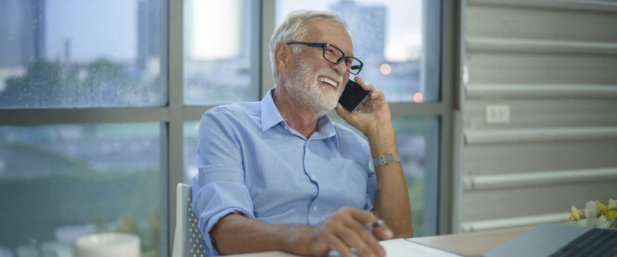 An older man sitting at a desk, engaged in a conversation on the phone. 