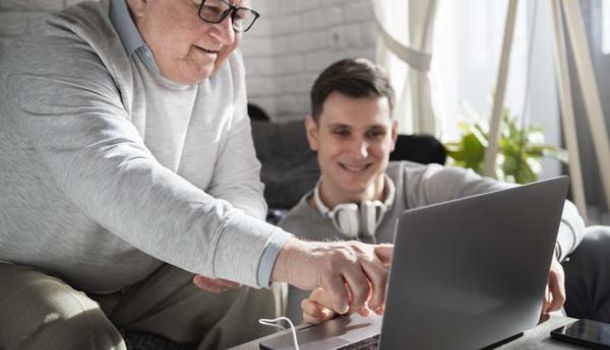 An older man and a younger man sit together on a couch, focused on a laptop in front of them.  