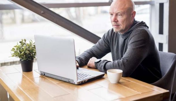 A bald man seated at a table, focused on his laptop, with a neutral background. 