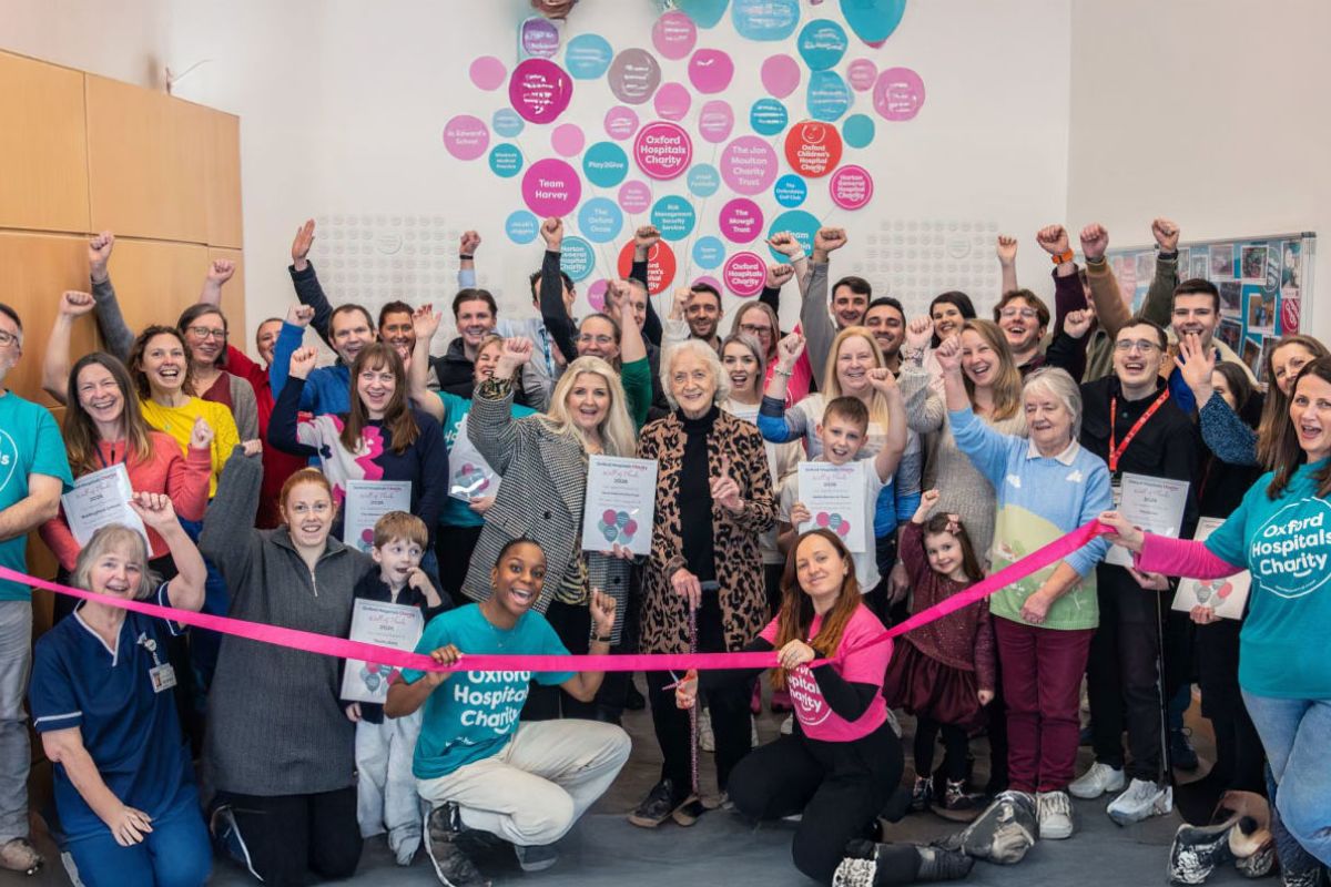 Group of Oxford Hospitals Charity staff, volunteers, supporters and community members celebrating together at a ribbon-cutting ceremony, with raised arms, certificates and a colourful donor recognition display on the wall behind them.