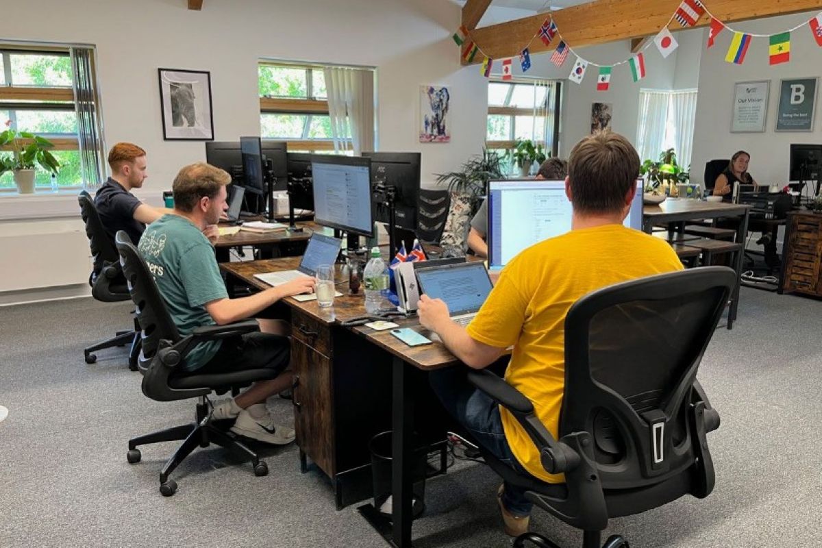 Open-plan office with a team of employees working at desktop computers in a bright modern workspace, featuring wooden ceiling beams, large windows, indoor plants, and international flags hanging overhead.