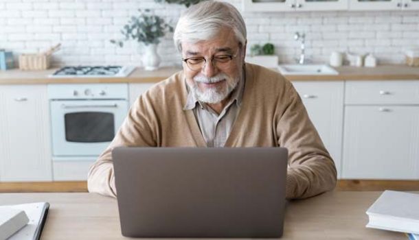 An older man sitting at a table, focused on his laptop, with a thoughtful expression on his face.  